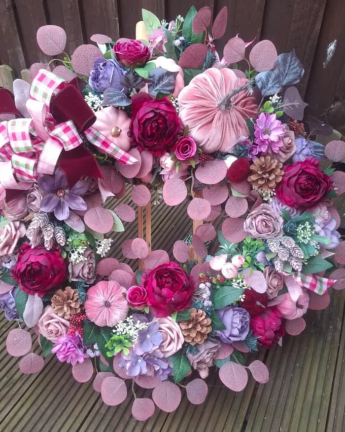 A wreath of pink, lilac and magenta blooms with gingham ribbon and frosted pinecones, on decking.