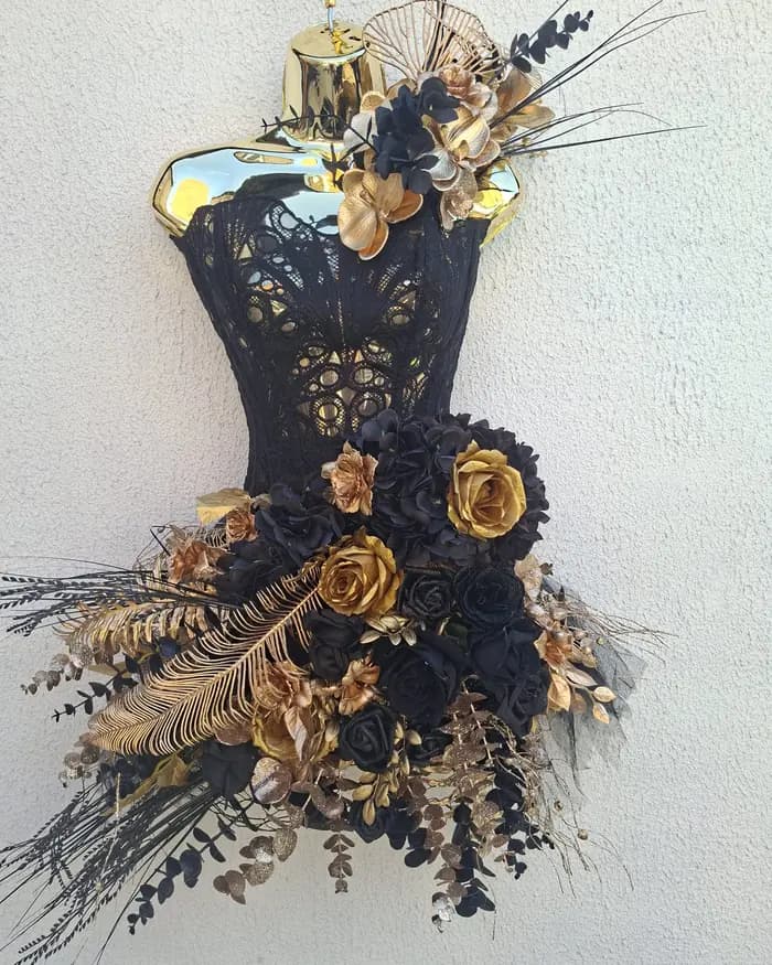 A black and gold corset-shaped wall piece with lace, gold roses and feathers, hung on a white rendered wall.