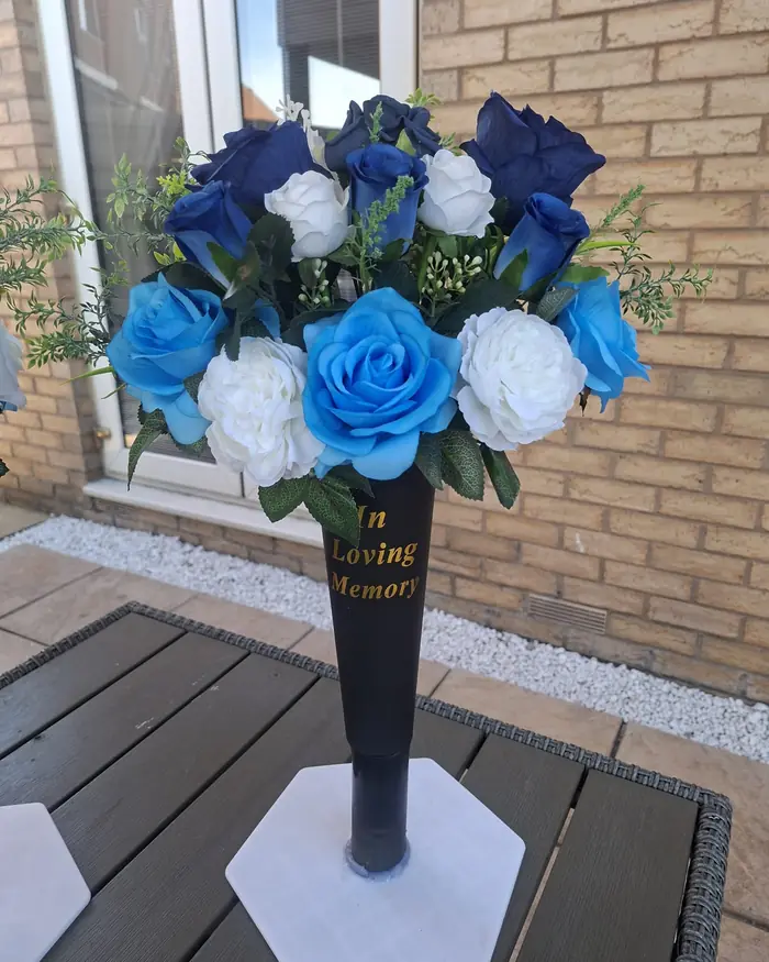 Blue and white roses in a black In Loving Memory graveside vase, against a brick wall.