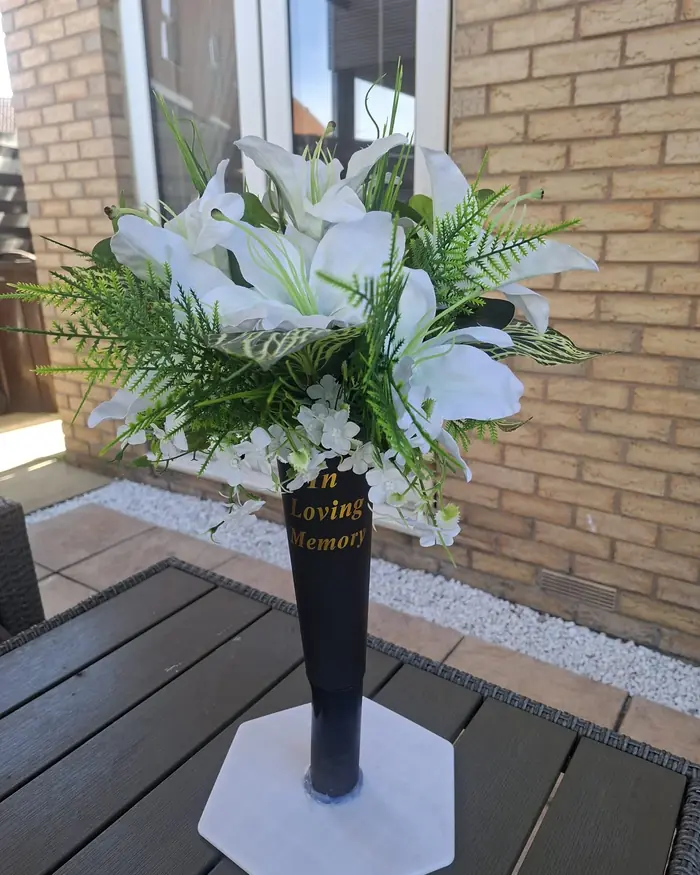 White lilies, white blossom and green ferns in a black In Loving Memory graveside vase.