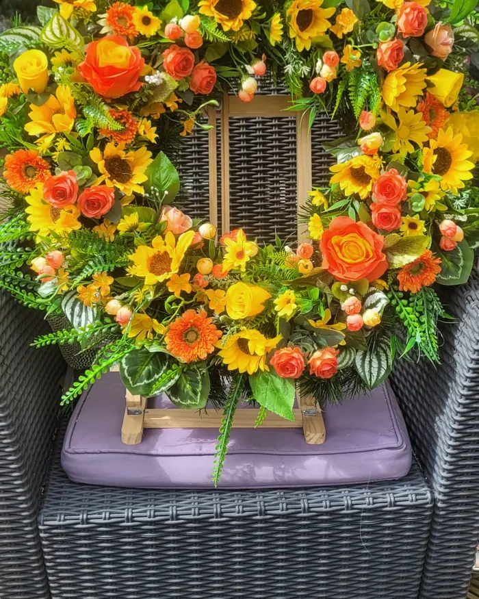 A yellow sunflower and orange rose wreath on a wooden easel, set on a garden chair.
