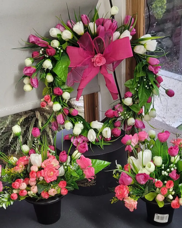 A pink and white rose wreath with a pink bow, with small potted arrangements beneath it.