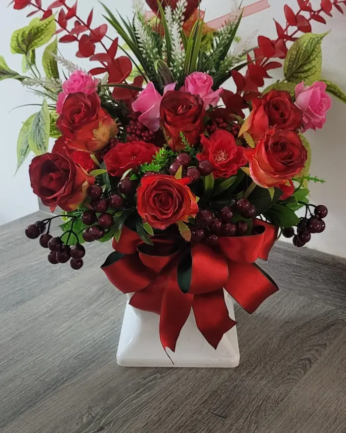 Red roses and berries finished with a red bow, in a white pot on a table.