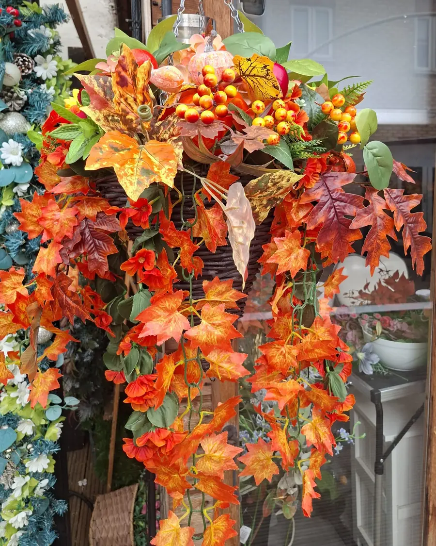 A dark wicker cone hanging basket trailing orange maple leaves with clusters of orange and yellow berries and a pale pink pumpkin at the top.
