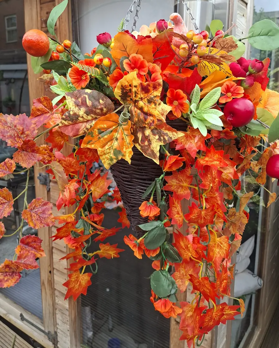 A dark wicker cone hanging basket with a long trail of orange and red maple leaves, berries, an apple and a small pumpkin, hanging beside a wooden framed window.