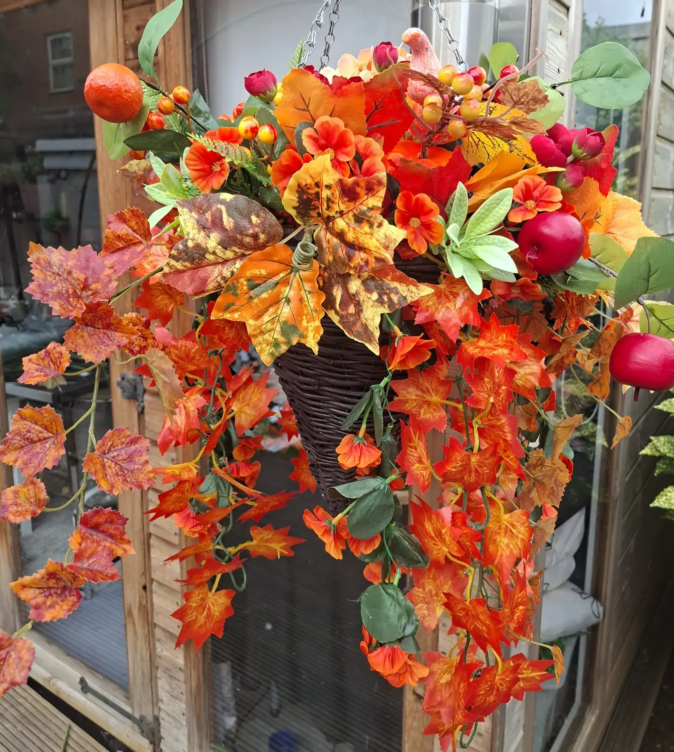 A dark wicker cone hanging basket with a long trail of orange and red maple leaves, berries, an apple and a small pumpkin, hanging beside a wooden framed window.