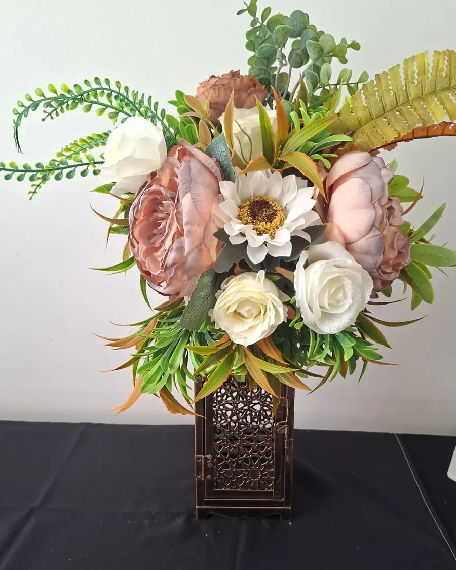 A copper fretwork lantern holding blush peonies, cream roses, a white sunflower and trailing green ferns.