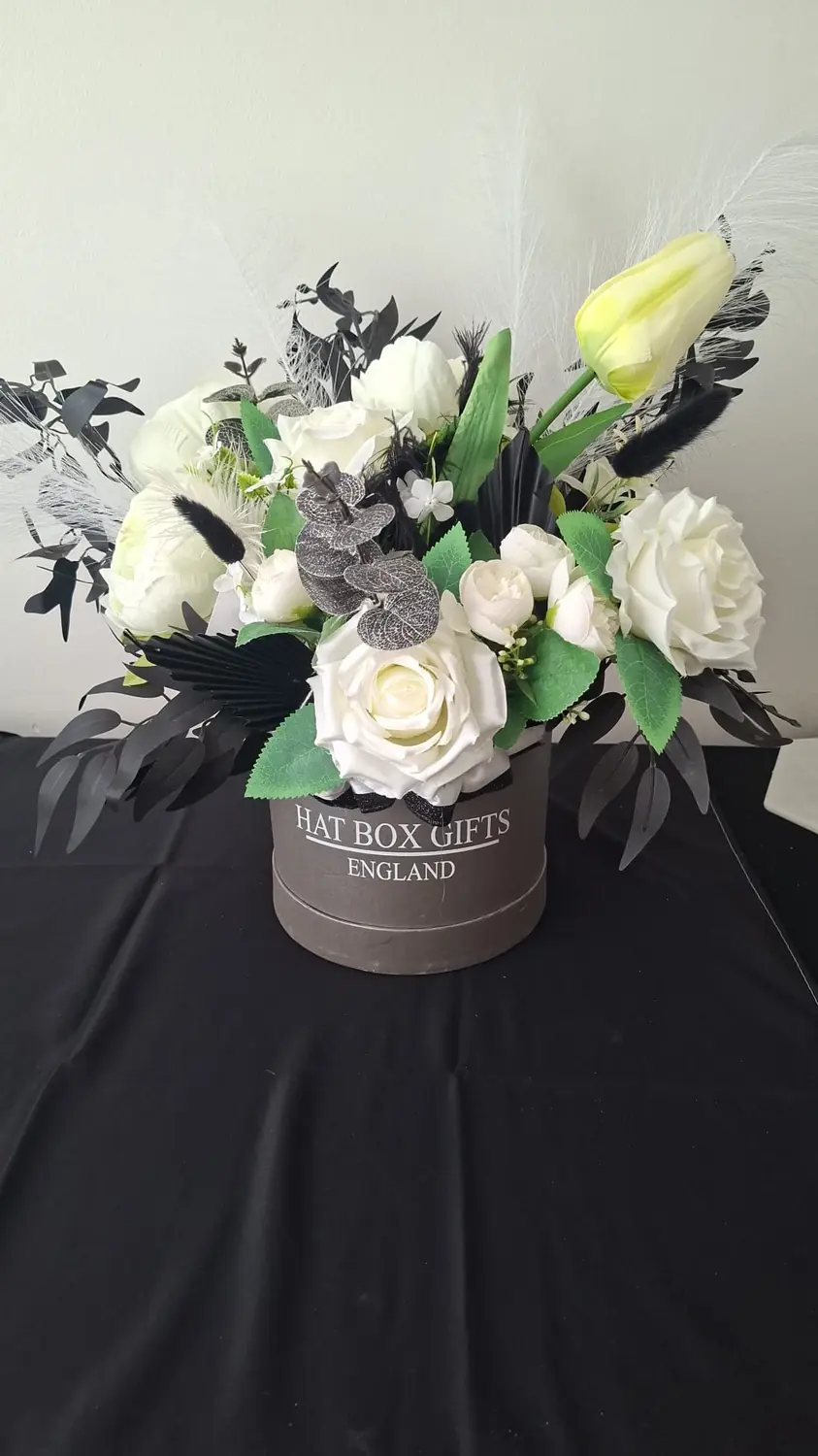 A grey hat box arrangement of white roses and tulips with black palm spears, black bunny tails and white pampas grass.