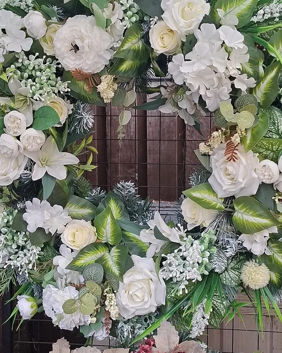 A large wreath of white roses, lilies and hydrangeas set in deep green variegated foliage, hung on a black metal display panel.