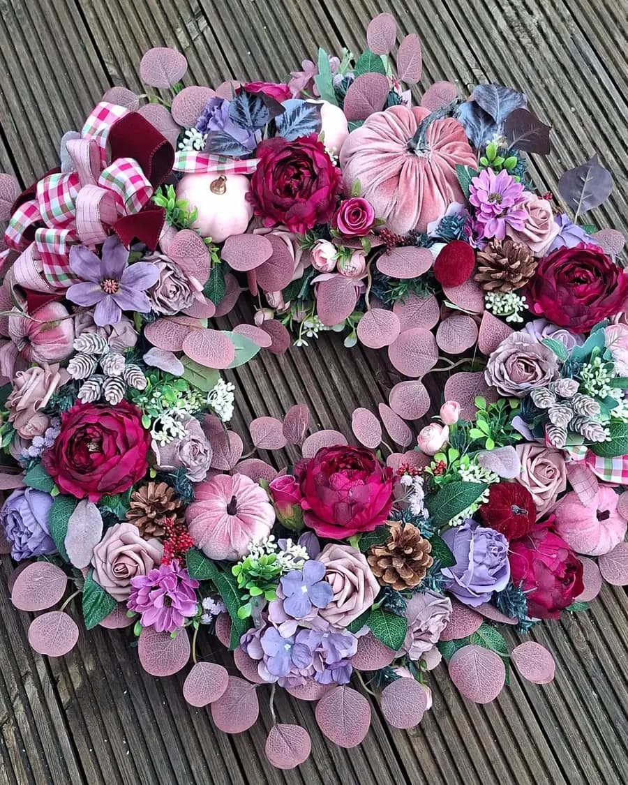 A 65cm autumn wreath in dusky pink, lilac and magenta, with velvet pumpkins, pinecones and pink gingham ribbon, laid flat on weathered grey decking.