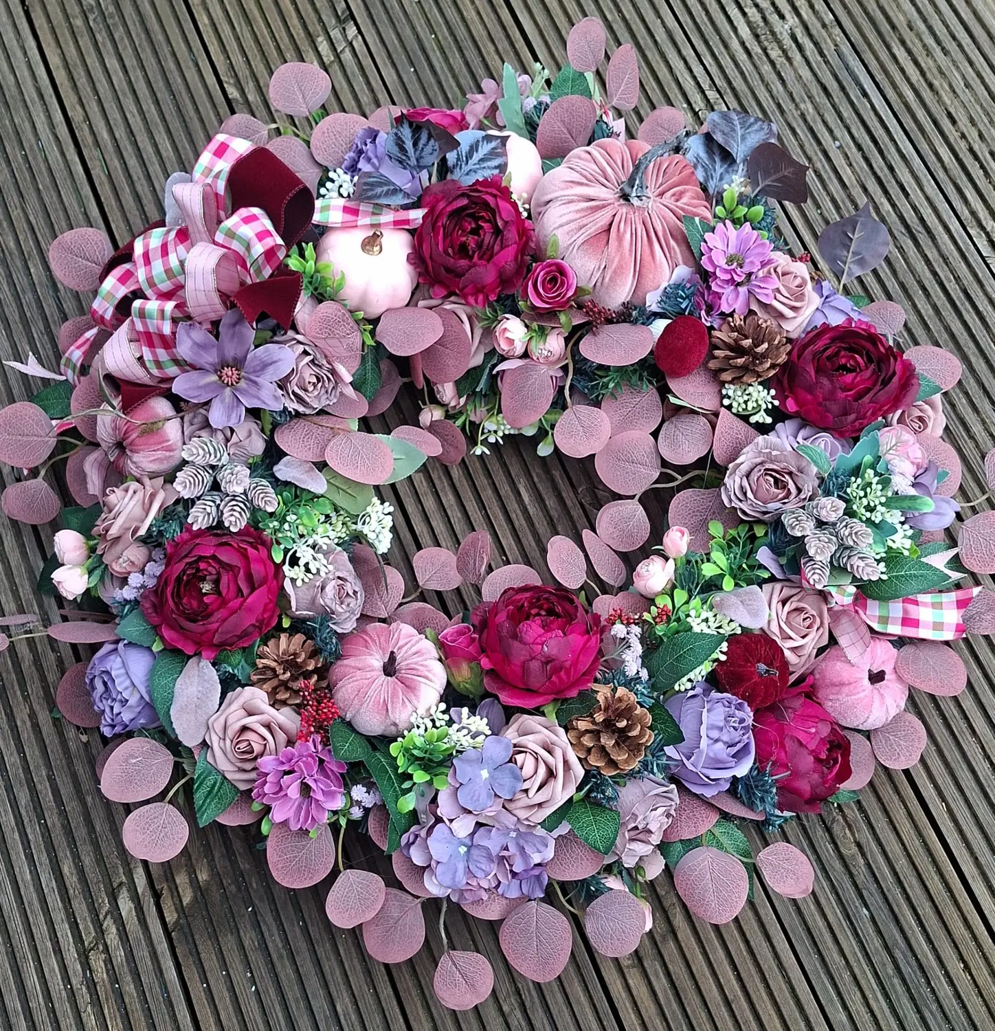 A 65cm autumn wreath in dusky pink, lilac and magenta with velvet pumpkins, laid flat on weathered grey decking.