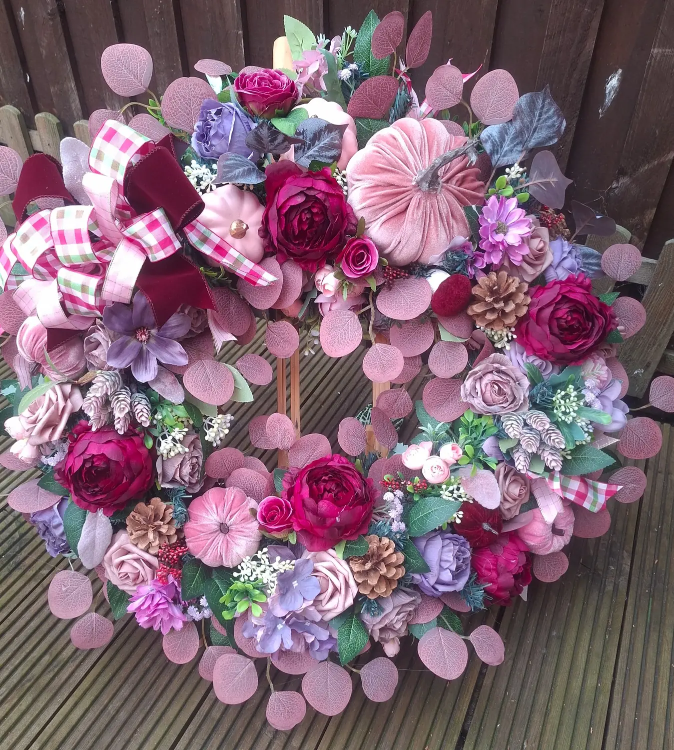 A wreath of pink, lilac and magenta blooms with gingham ribbon and frosted pinecones, on decking.