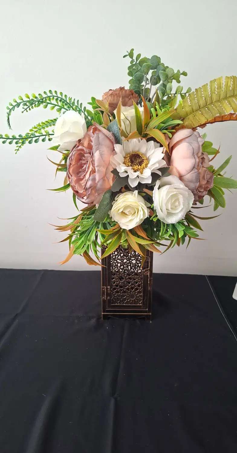 A copper fretwork lantern holding blush peonies, cream roses, a white sunflower and trailing green ferns.
