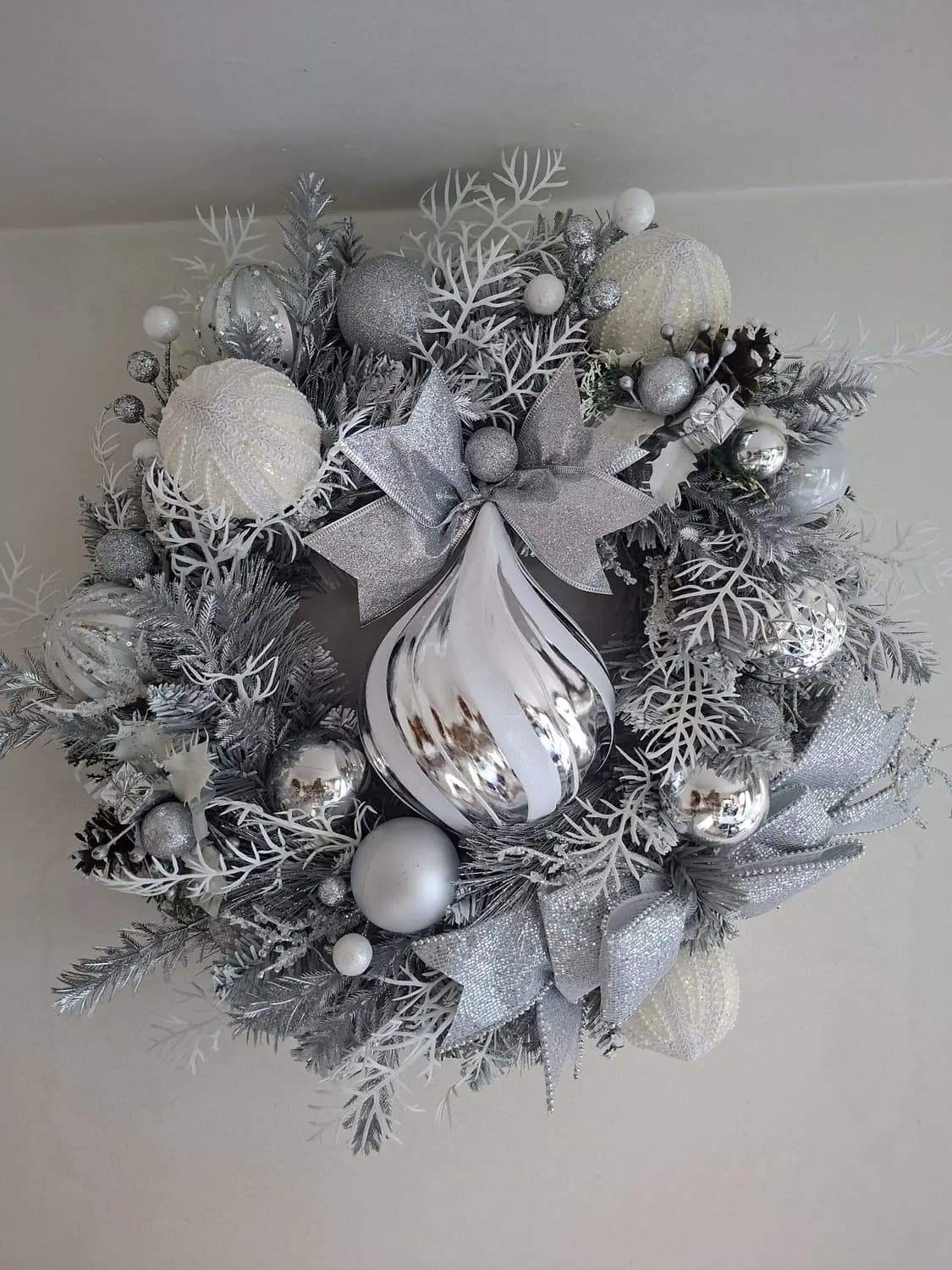 A Christmas wreath of frosted silver and white foliage, silver and pearl baubles and a glitter bow, with a large silver teardrop bauble hanging at its centre.